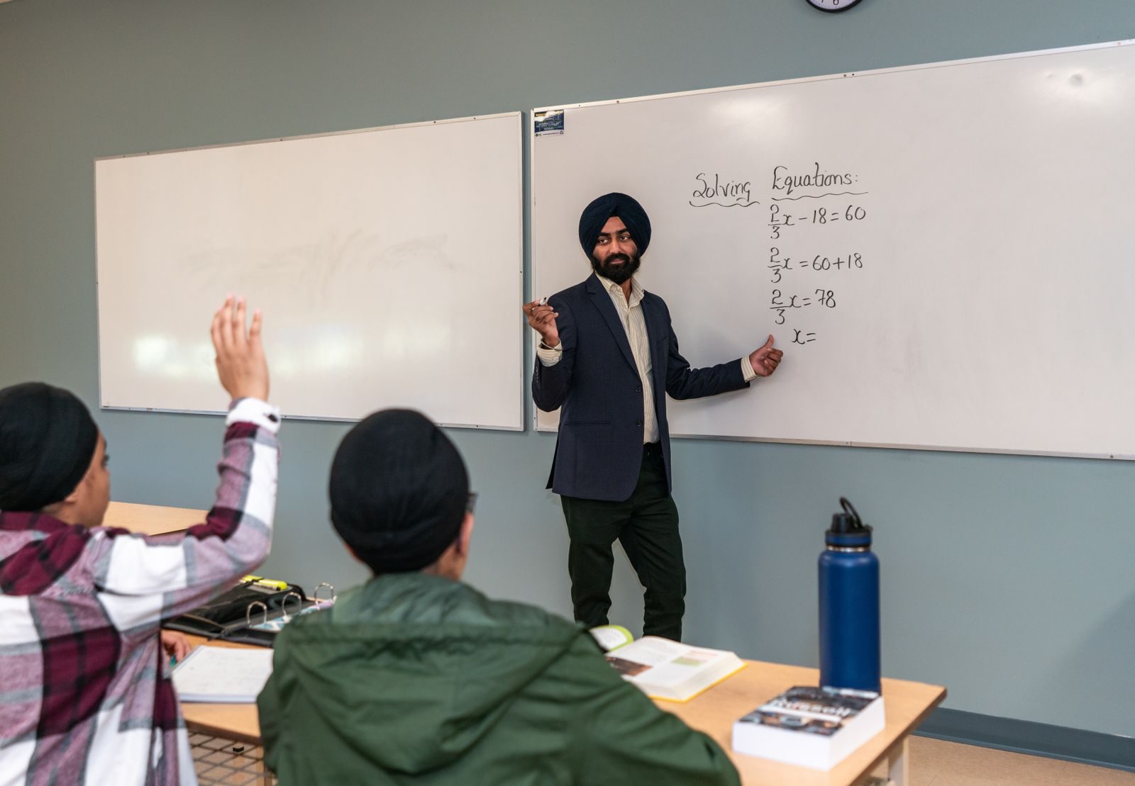 A maths teacher taking a class at the whiteboard while a student raises a hand